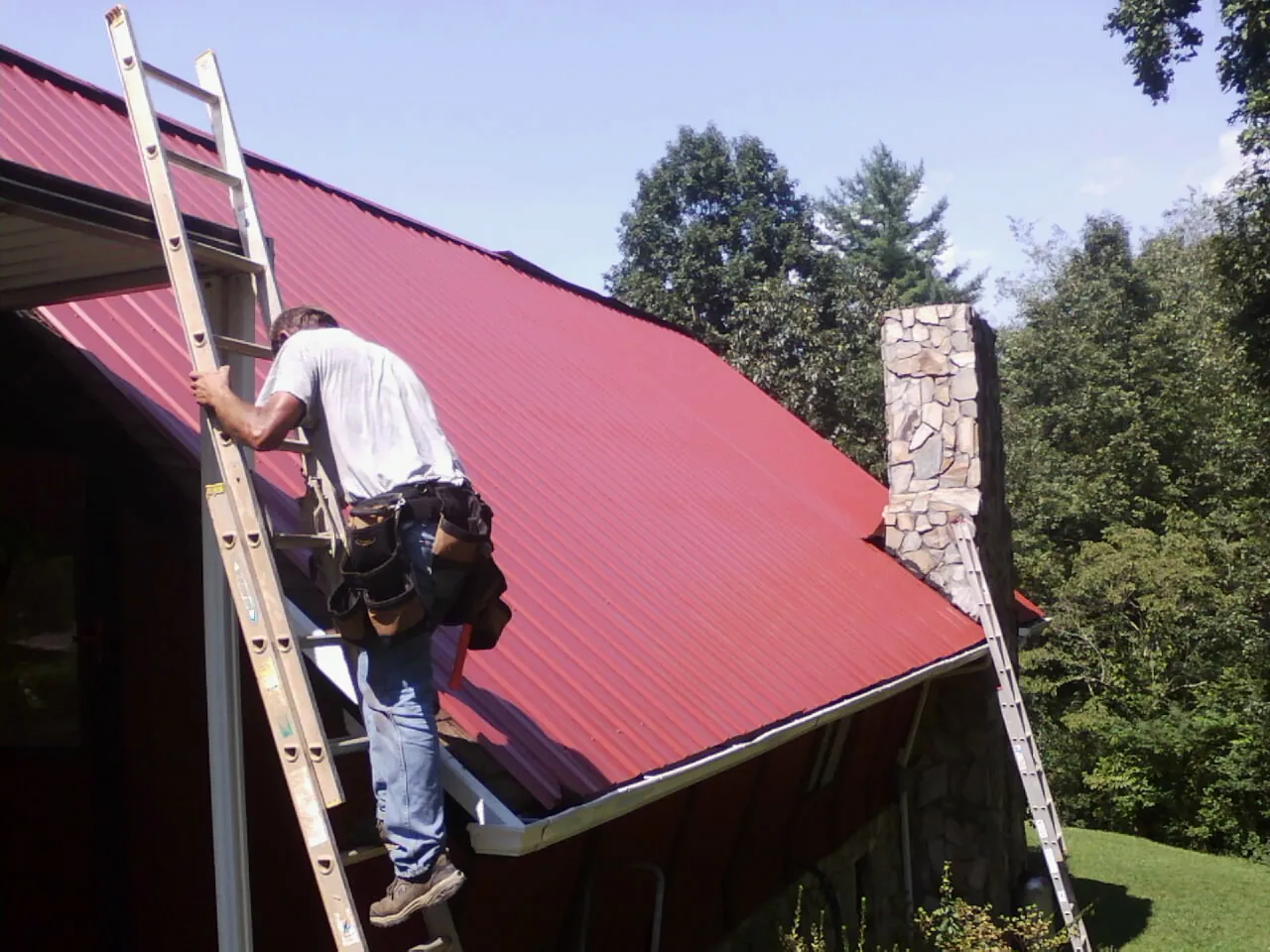 Roofer on a ladder beside a red metal roof and stone chimney — Soffit & Fascia Repair by Roofing Brothers in Ashley